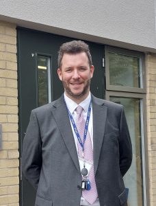 Smiling man in a gray suit and pink tie, wearing a staff lanyard outside a building entrance.